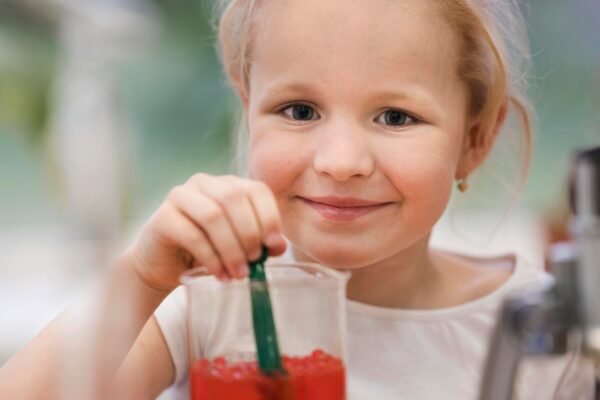 Young toddler holding a sippy cup of cranberry juice while sitting at a table with a watchful parent.